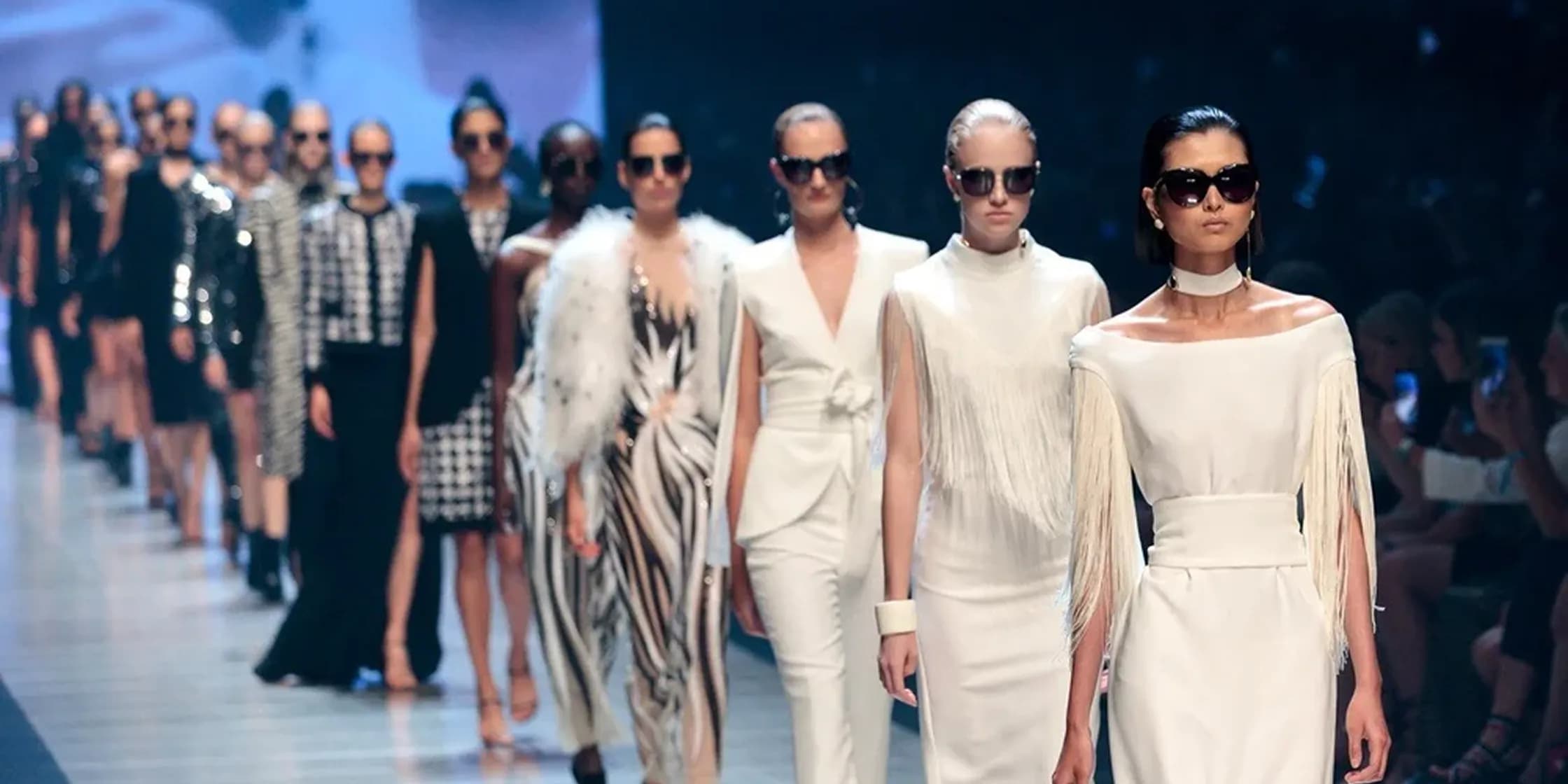 A line of models walking a runway during a fashion show, wearing looks ranging from patterned to all-white ensembles
