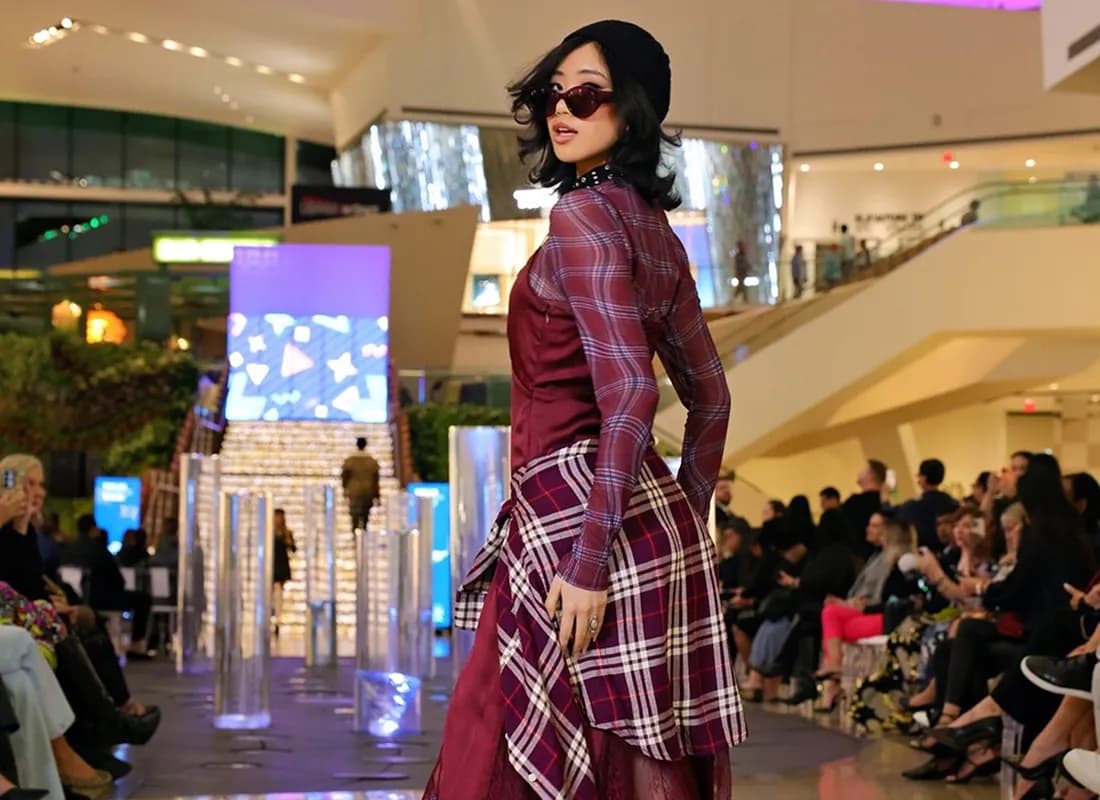 A model presenting a layered plaid and burgundy look on a runway in a shopping center atrium